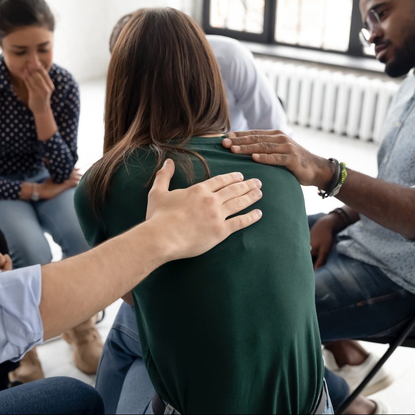Multi ethnic people gathered together sitting on chairs in circle supporting crying desperate girl during group therapy session, medical detox center, psychological help assistance at meeting concept